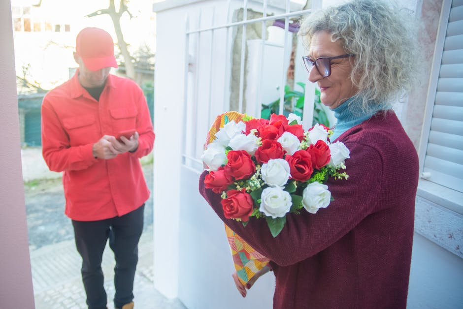 A woman holding a vibrant bouquet of fresh roses, featuring red and white blooms with green foliage, wrapped in colorful decorative paper, stands at her doorway. She is wearing glasses, a maroon sweat