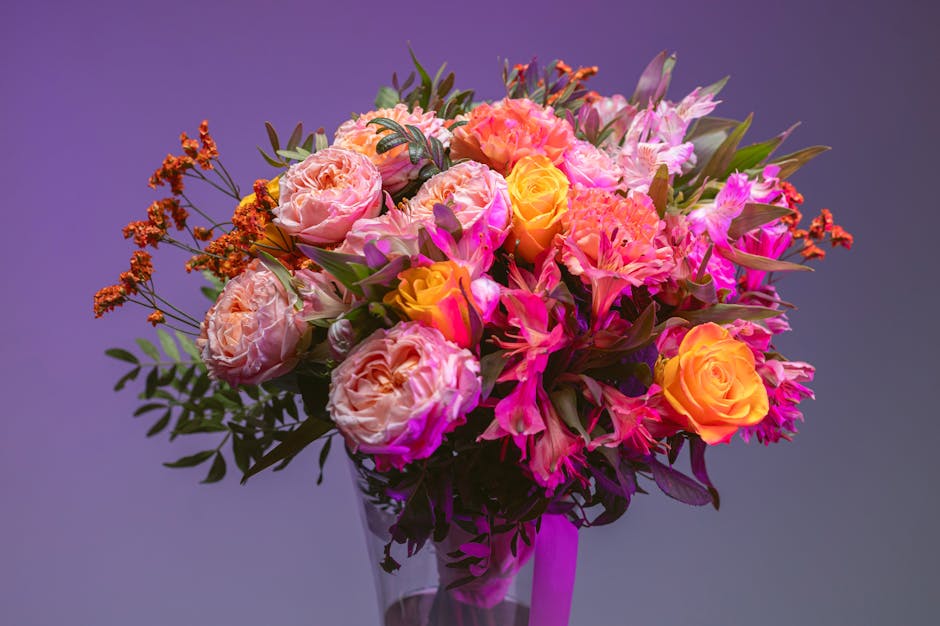 A vibrant floral bouquet featuring large, soft pink peonies, bright yellow roses, and striking pink orchids arranged in a clear glass vase. The arrangement includes lush green foliage and delicate red