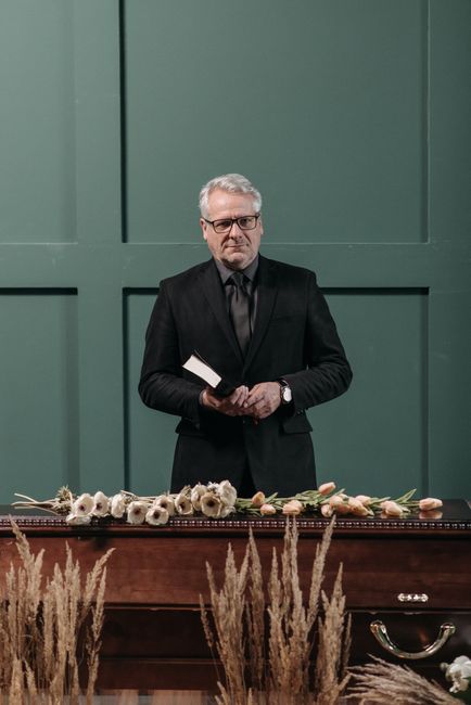 A man dressed in a black suit and glasses stands behind a wooden table, holding a small white book or card in his hands. In front of him, on the table, there is a floral arrangement consisting of whit