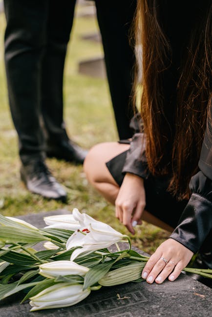 A floral arrangement consisting of white lilies with large, smooth petals and prominent stamens, arranged on a dark stone surface. The lilies are surrounded by long, green leaves with a glossy finish.