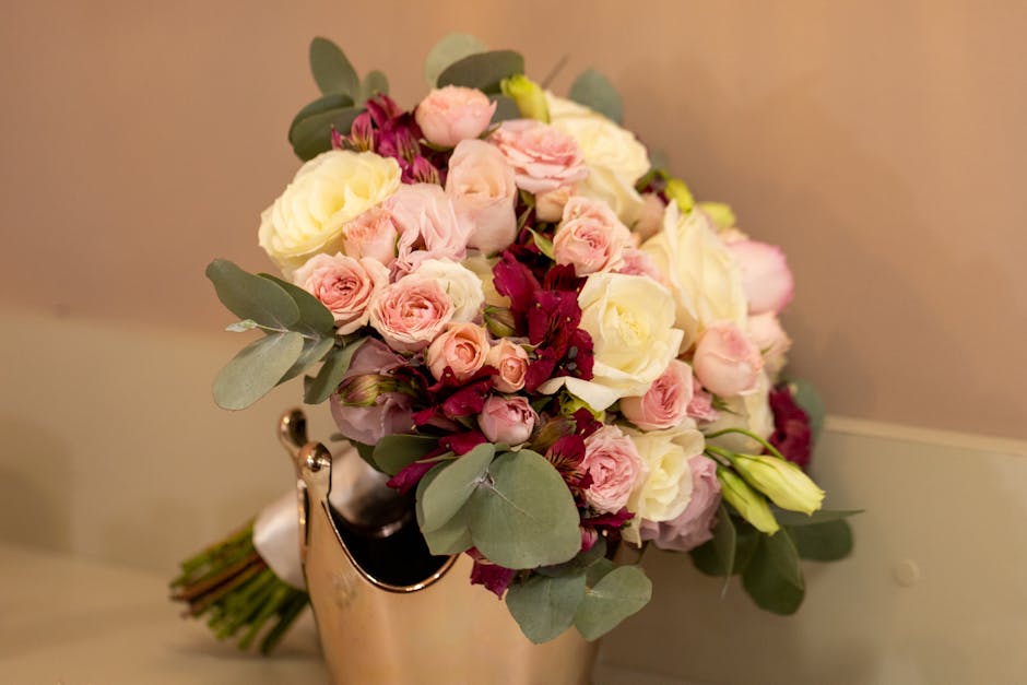A bouquet of fresh flowers featuring cream and pale pink roses, complemented by deep burgundy blooms and dusty green eucalyptus leaves. The arrangement is gathered tightly in a rounded style and place