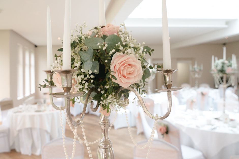 A close-up of a floral arrangement featuring soft pink roses, white baby's breath, and lush greenery, displayed in a silver candelabrum adorned with hanging pearl garlands. The arrangement is set in a