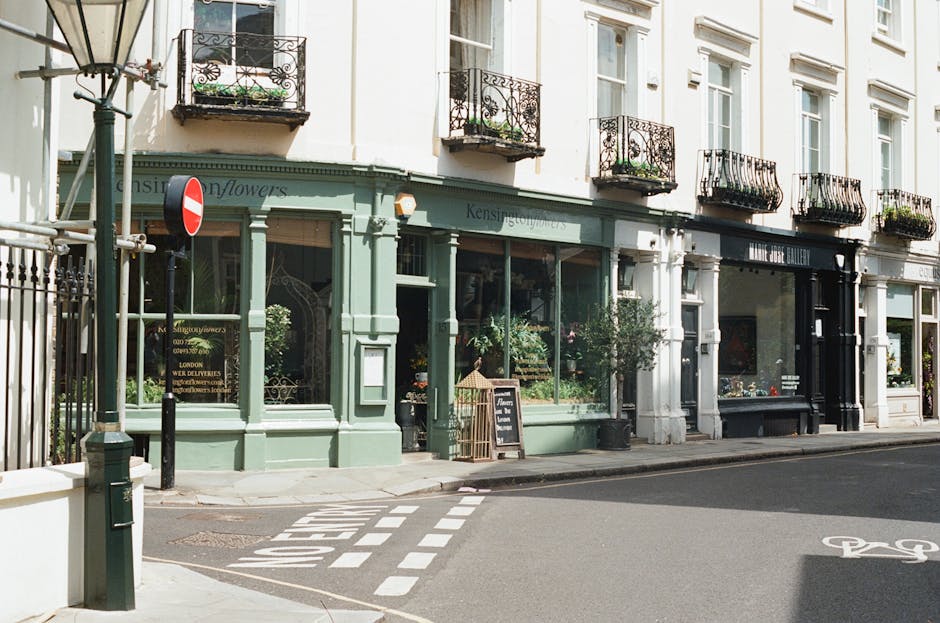 A charming flower shop with a sage green exterior and large glass windows displaying various floral arrangements. The shop features a small blackboard sign on the sidewalk and potted plants near the e